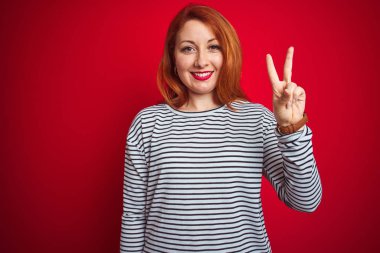 Genç kızıl saçlı kadın kırmızı izole arka plan üzerinde duran kayışlar lacivert gömlek giyen gösteren ve emin ve mutlu gülümserken parmakları ile iki işaret.
