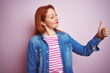 Denim gömlek giyen güzel kızıl saçlı kadın ve izole pembe arka plan üzerinde çizgili t-shirt gururlu arıyorsunuz, yan jest kadar başparmak yapıyor gülümseyerek