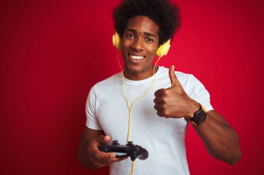 Afro Gamer adam büyük bir gülümseme ile izole kırmızı arka plan üzerinde joystick kulaklıklar kullanarak video oyunu oynarken ok işareti yapıyor, parmaklar ile başparmak, mükemmel işareti