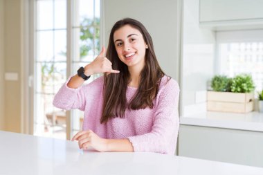 Pembe kazak giyen güzel genç kadın telefonda konuşmak gibi el ve parmaklarıyla telefon hareketi yaparak gülümsüyor. İletişim kavramları.