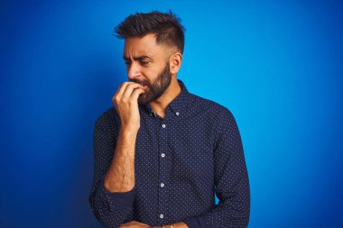 Genç Hint zarif adam örtü giyen gömlek ayakta izole mavi arka plan görünümlü vurguladı ve gergin elleri ile ağız ısırma çivi. Anksiyete sorunu.
