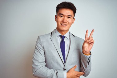 Asya Çinli işadamı giyen ve kravat ayakta izole beyaz arka plan ile gülümseyen mutlu yüz kırmızı kamera zafer işareti yapıyor. İki numara.