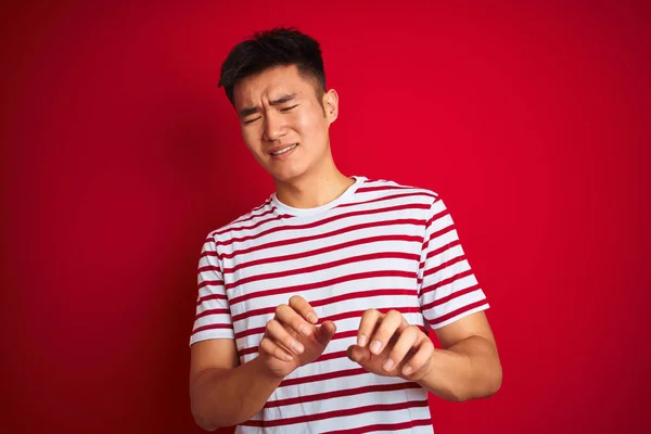 Young asian chinese man wearing striped t-shirt standing over isolated red background disgusted expression, displeased and fearful doing disgust face because aversion reaction. With hands raised. Annoying concept.