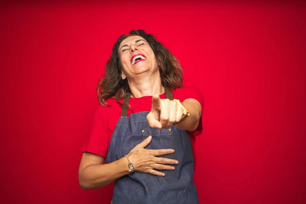 Middle age senior woman wearing apron uniform over red isolated background laughing at you, pointing finger to the camera with hand over body, shame expression