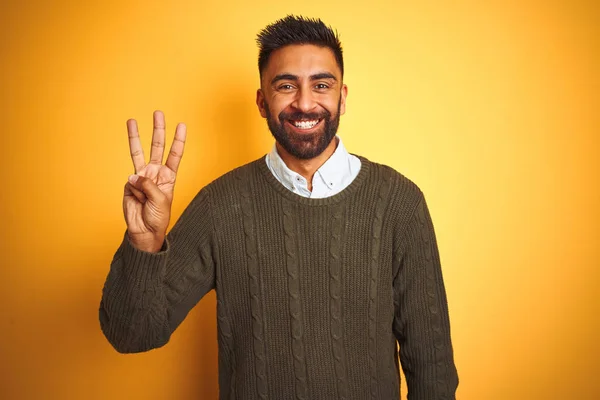 緑色のセーターとシャツを着た若いインド人男性が 自信を持って幸せに微笑みながら 3番の指を見せ 指で指を上げている黄色の背景の上に立っている — ストック写真