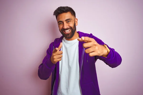 Young indian man wearing purple sweatshirt standing over isolated pink background pointing fingers to camera with happy and funny face. Good energy and vibes.