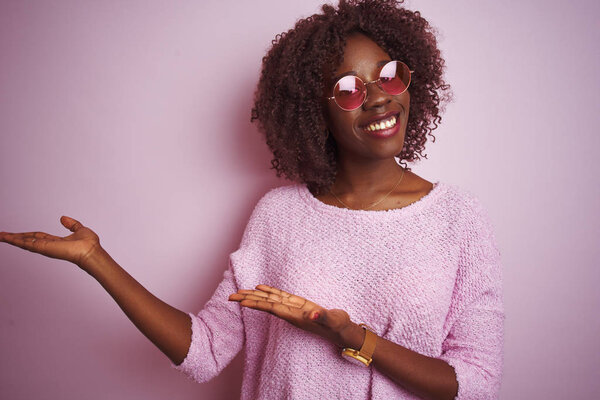 Young african afro woman wearing sweater and sunglasses over isolated pink background amazed and smiling to the camera while presenting with hand and pointing with finger.