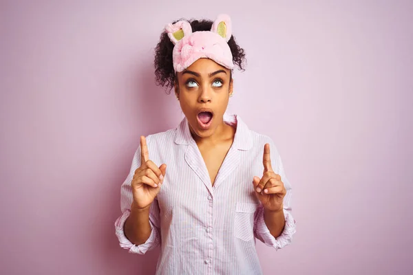 年轻的非洲裔美国妇女穿着睡衣和面具在孤立的粉红色背景惊讶和惊讶抬头 用手指和抬起手臂 — 图库照片