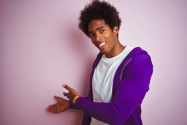 孤立したピンクの背景の上に立つ紫色のスウェットシャツを着た若いアフリカ系アメリカ人男性が 開いた手で自然に微笑む — ストック写真