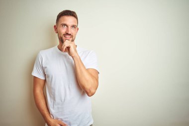 Genç yakışıklı adam soru, dalgın ifade hakkında düşünme çene üzerinde el ile izole arka plan üzerinde rahat beyaz t-shirt giyiyor. Gülümseyen ve düşünceli bir yüz. Şüphe kavramı.