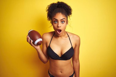 Tatilde bikini giyen afro-amerikalı kadın izole sarı arka plan üzerinde rugby oynarken sürpriz bir yüz ile şok korkmuş, korku ve korku ifade ile heyecanlı