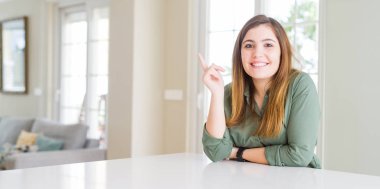 Kameraya bakarak yan el ve parmak ile işaret eden güzel genç kadın evde yüzünde kocaman bir gülümseme ile.