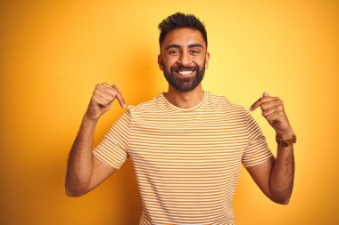Genç Hint adam giyen t-shirt ayakta izole sarı arka plan üzerinde yüz gülümseme ile emin görünümlü, parmaklarıyla gurur ve mutlu kendini işaret.