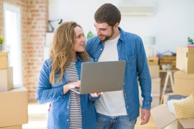 Yeni bir daireye taşımak için mutlu karton kutular etrafında bir odada bilgisayar laptop ayakta kullanarak genç çift