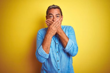 İzole sarı arka plan üzerinde duran denim gömlek giyen yakışıklı orta yaş adam hata için elleri ile ağız kapsayan şok. Gizli kavram.