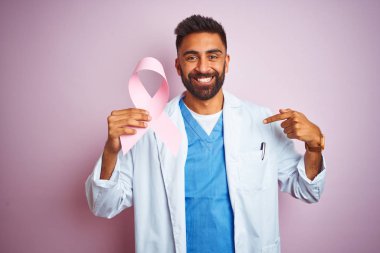 Genç Hintli doktor adam kendine parmak işaret sürpriz yüzü ile izole pembe arka plan üzerinde duran kanser kurdele tutan