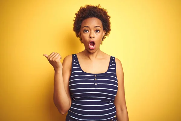 Beauitul african american woman wearing summer t-shirt over isolated yellow background Surprised pointing with hand finger to the side, open mouth amazed expression.