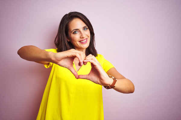 Молодая красивая женщина в желтой футболке стоит на розовом изолированном фоне улыбаясь в любви показывая символ сердца и форму с руками. Романтическая концепция
.