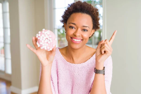 Young african american woman eating pink sugar marshmallows donut very happy pointing with hand and finger to the side
