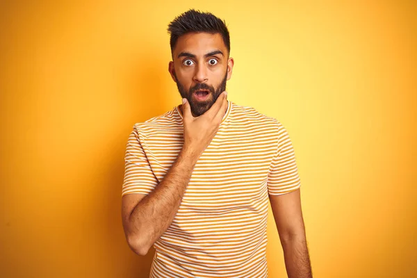 Young indian man wearing t-shirt standing over isolated yellow background Looking fascinated with disbelief, surprise and amazed expression with hands on chin