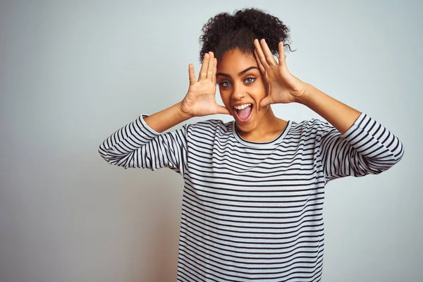 African american woman wearing navy striped t-shirt standing over isolated white background Smiling cheerful playing peek a boo with hands showing face. Surprised and exited
