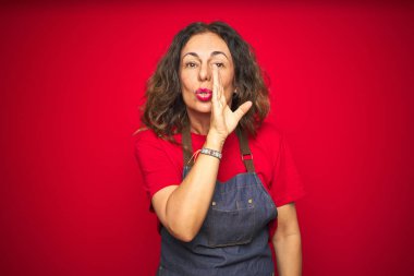 Orta yaş yaşlı kadın ağız üzerinde kırmızı izole arka plan el üzerinde önlük üniforma giyen gizli söylenti söylüyorum, kötü niyetli konuşma konuşma fısıldıyor