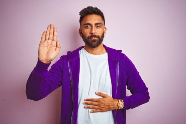 Mor sweatshirt giyen genç hintli adam göğüs ve açık avuç içi el ile izole pembe arka plan üzerinde duran bir sadakat sözü yemini yapma
