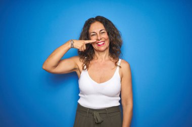 Kıvırcık saçlı orta yaş yaşlı kadın mavi izole arka plan üzerinde duran yüz ve burun el parmağı ile işaret, neşeli gülümseyen. Güzellik konsepti