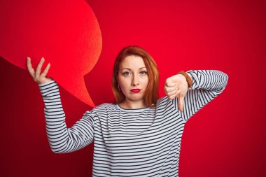 Genç güzel kızıl saçlı kadın kızgın yüz ile kırmızı izole arka plan üzerinde konuşma balonu tutan, başparmak aşağı, ret kavramı ile sevmediğimi gösteren negatif işareti