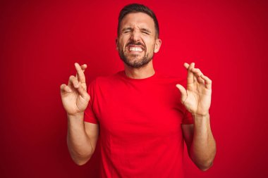 Kırmızı izole arka plan üzerinde rahat t-shirt giyen genç yakışıklı adam umut ve gözler ile gülümseyen geçti parmak kapalı. Şans ve batıl fikir.