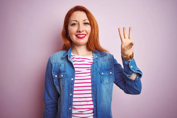 デニムシャツとストライプのTシャツを着た美しい赤毛の女性は 自信を持って幸せに微笑みながら 指番号3を示し 指で指を上げている — ストック写真