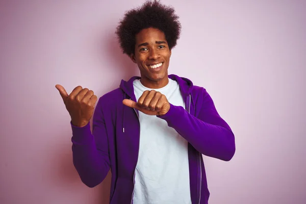 孤立したピンクの背景の上に立つ紫色のスウェットシャツを着た若いアフリカ系アメリカ人男性は 手と親指で後ろを指し 自信を持って微笑んでいる — ストック写真