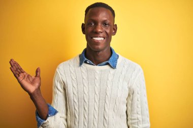 Kot gömlek ve beyaz kazak giyen Afrikalı Amerikalı adam izole sarı arka plan gülümseyen neşeli sunan ve kamera bakarak el avuç ile işaret.
