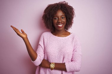 Genç Afrikalı Afro kadın izole pembe arka plan gülümseyen neşeli sunumu üzerinde ayakta kazak giyen ve kamera bakarak el avuç ile işaret.