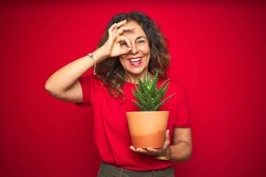 Orta yaş üst düzey kadın kırmızı izole arka plan üzerine yeşil kaktüs tutan mutlu yüz gözleri parmak görünümlü el ile ok işareti yapıyor gülümseyen