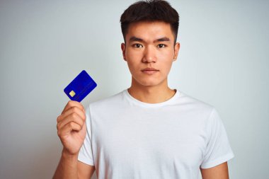 Young asian chinese man holding credit card standing over isolated white background with a confident expression on smart face thinking serious clipart