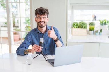 Genç adam kayıt podcast kullanarak mikrofon ve laptop mutlu büyük gülümseme yapıyor ok işareti, başparmak yukarı parmaklar, mükemmel işareti