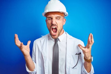 Genç yakışıklı mühendis adam mavi izole arka plan üzerinde emniyet kask giyen deli ve silah ile başarı için deli kutluyor kaldırdı ve heyecanlı çığlık kapalı gözleri. Kazanan kavramı