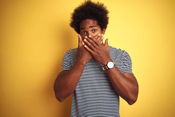 African american man with afro hair wearing navy striped t-shirt over isolated yellow background shocked covering mouth with hands for mistake. Secret concept.