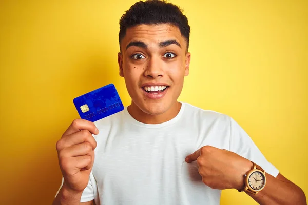 年轻的巴西客户男子拿着信用卡站在孤立的黄色背景与惊喜的脸指向自己 — 图库照片