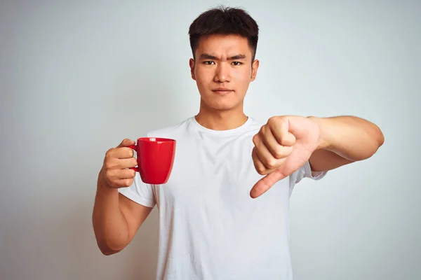 Asian chinese man holding red cup of coffee standing over isolated white background with angry face, negative sign showing dislike with thumbs down, rejection concept