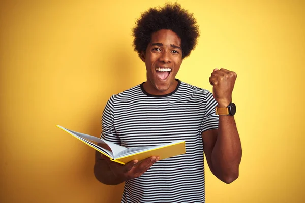 Afro american student man reading book standing over isolated yellow background screaming proud and celebrating victory and success very excited, cheering emotion