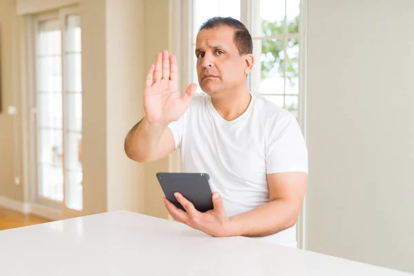 Middle age man using digital table at home with open hand doing stop sign with serious and confident expression, defense gesture