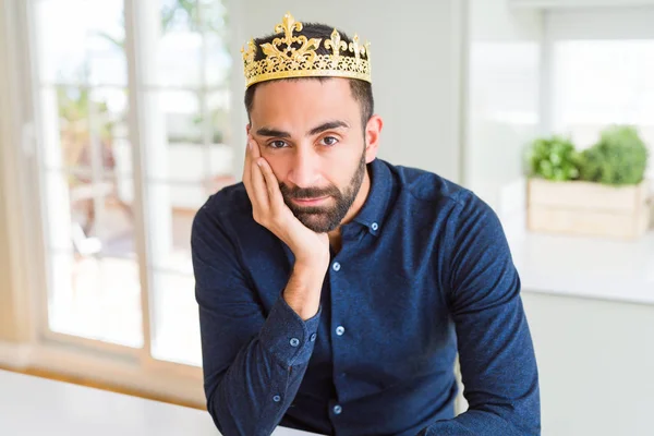 Handsome hispanic man wearing golden crown over head as the king thinking looking tired and bored with depression problems with crossed arms.