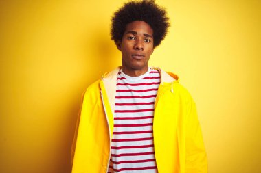Afro saç ile Afrikalı Amerikan adam izole sarı arka plan üzerinde ayakta yağmur ceket giyiyor yüzünde ciddi bir ifade ile rahat. Basit ve doğal kamera bakıyor.