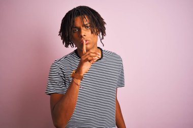 Zencili pembe arka plan üzerinde ayakta lacivert çizgili t-shirt giyen dreadlocks ile Afro adam dudaklarında parmak ile sessiz olmak istiyor. Sessizlik ve gizli konsept.