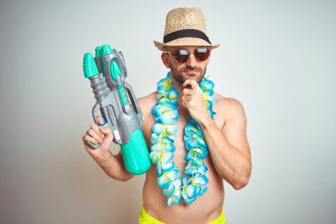 Yaz şapkası giyen genç adam ve Hawaii Lei çiçekler izole arka plan ciddi yüz hakkında soru düşünme üzerinde su tabancası tutan, çok karışık fikir