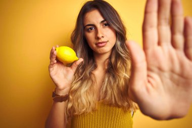 Açık el ile sarı izole arka plan üzerinde bir limon yeme genç güzel kadın ciddi ve emin ifade ile stop işareti yapıyor, savunma jest
