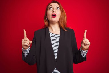 Güzel Redhead iş kadın izole kırmızı arka plan üzerinde zarif ceket giyiyor şaşırdı ve görünümlü ve parmaklar ve yükseltilmiş kollar ile işaret şaşırttı.