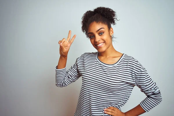 African american woman wearing navy striped t-shirt standing over isolated white background smiling looking to the camera showing fingers doing victory sign. Number two.